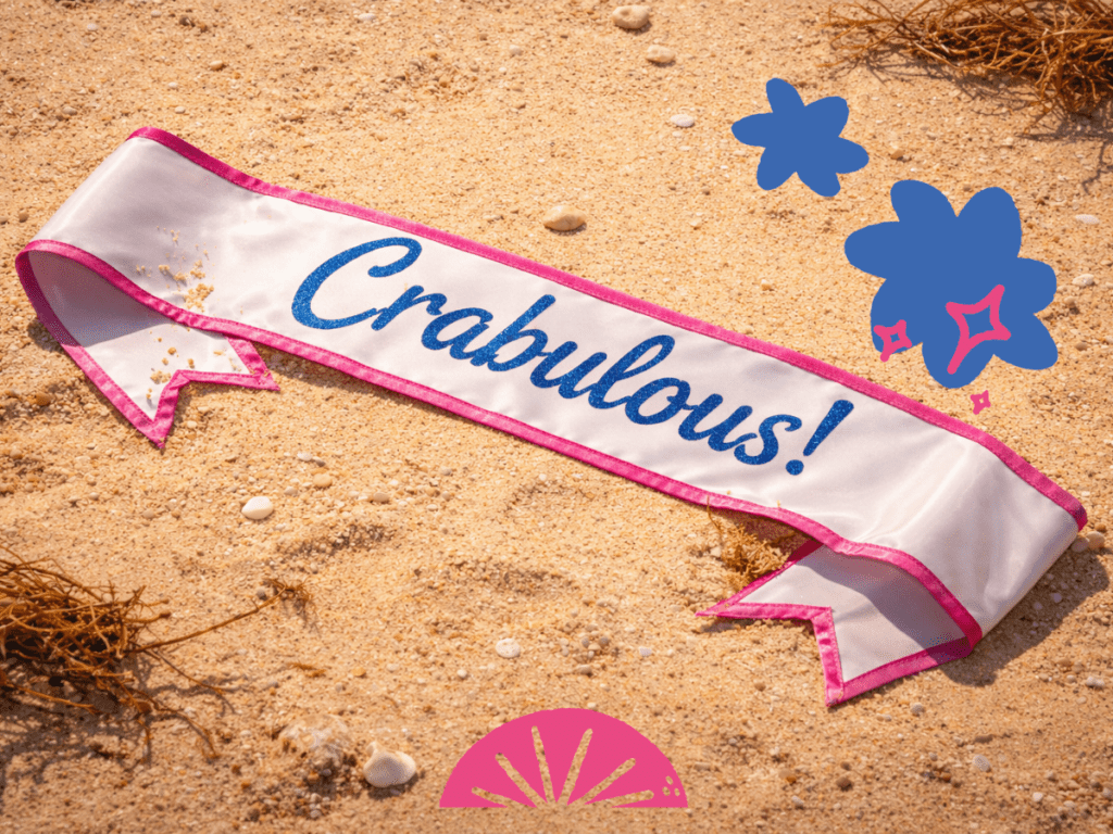 A pageant sash on a sandy beach that reads 'Crabulous!' in sparkly blue writing. 