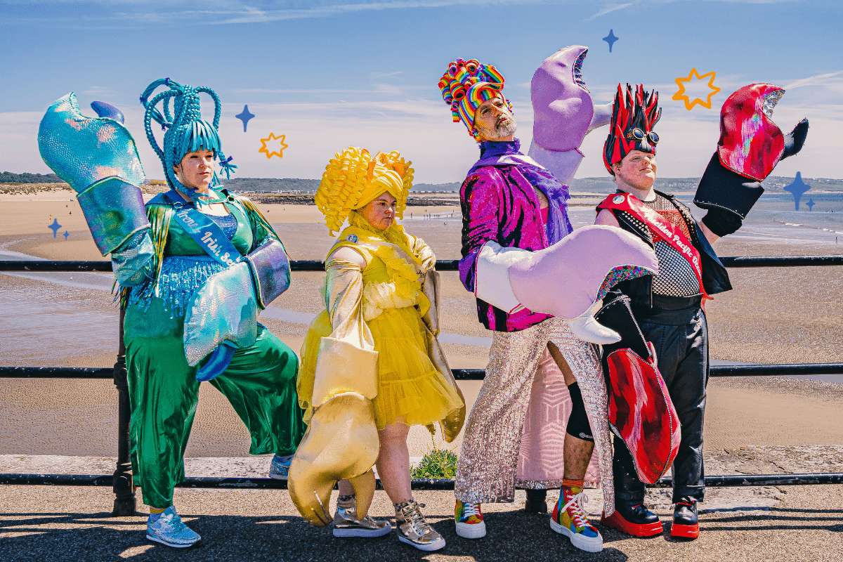 http://The%20four%20performers%20in%20their%20colourful%20crab%20costumes%20with%20huge%20crab%20pincers,%20stood%20posing%20next%20to%20railings%20by%20a%20beach.